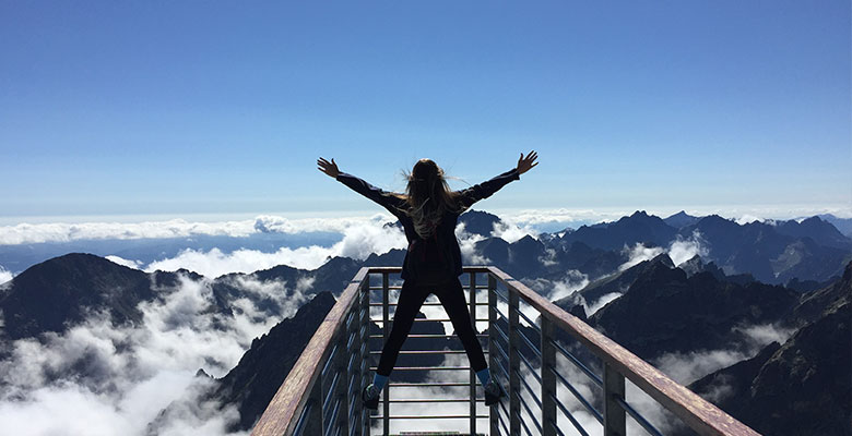 personne de dos, jambe écartées, en équilibre sur un parapet devant des sommets montagneux baignés de nuages